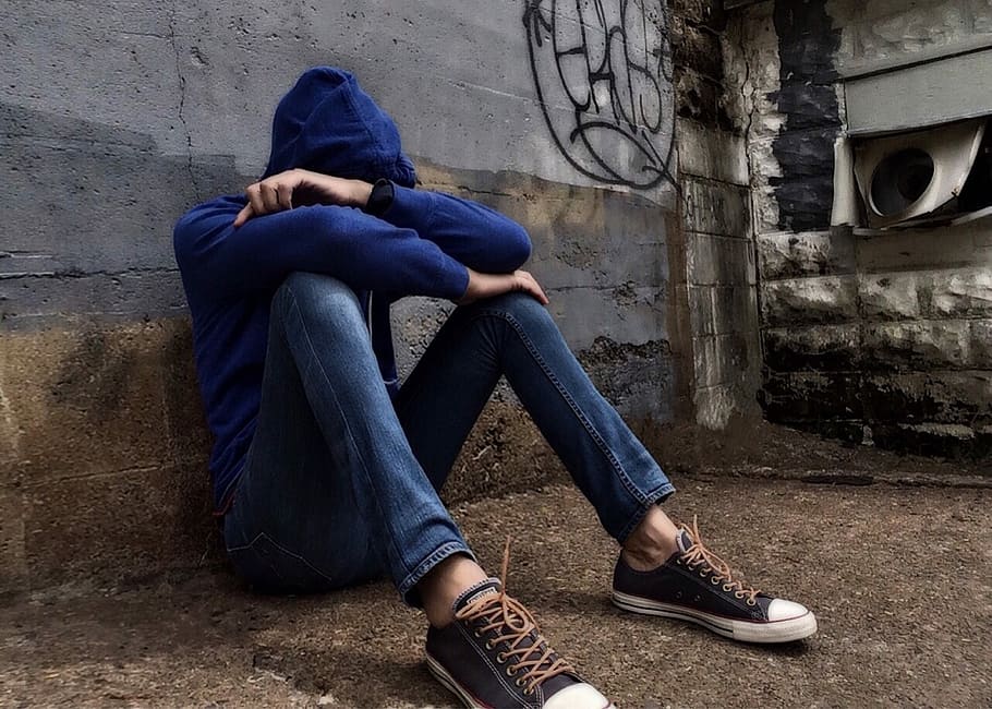 person, blue, pullover hoodie, jeans, sitting, leaning, gray, concrete, wall, daytime