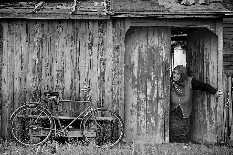 grayscale photo, woman opening, door, watching, bicycle, old, barn, bike, wooden house, old cottage