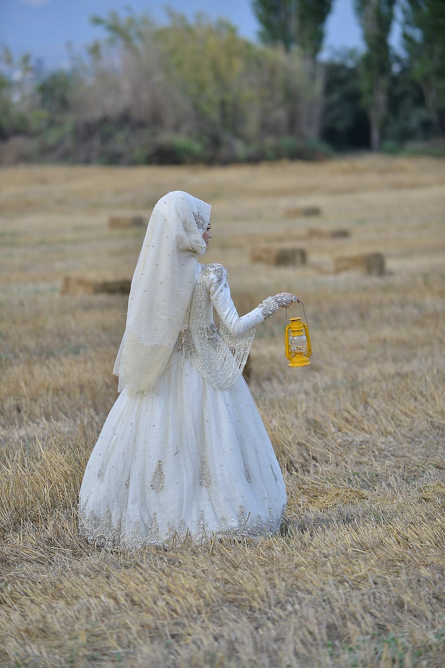 woman, white, wedding abaya, holding, yellow, kerosene lamp, brown, field, daytime, woman in white