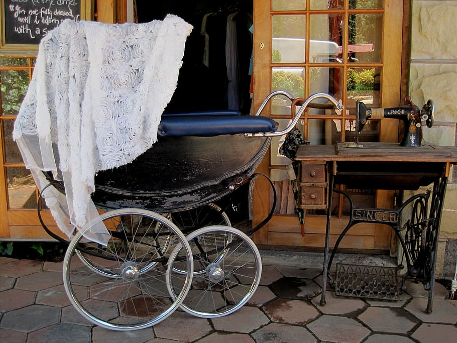 pram stroller, threadle sewing machine, black perambulator, large wheels, old fashioned, old sewing machine, white lace covering, grey floor paving, shop entrance, glass pane door