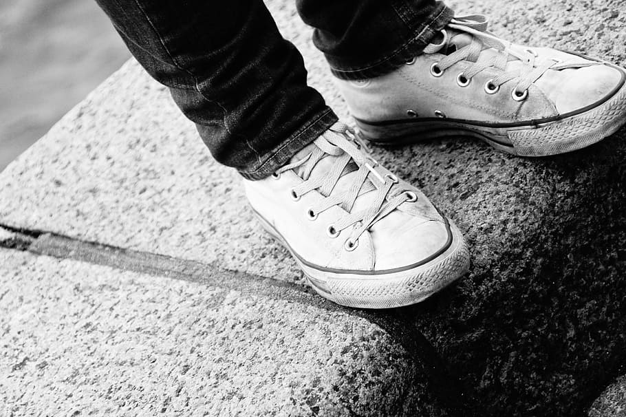 grayscale photography, standing, person, wearing, jeans, lace-up shoes, black and white, shoes, converse, black white