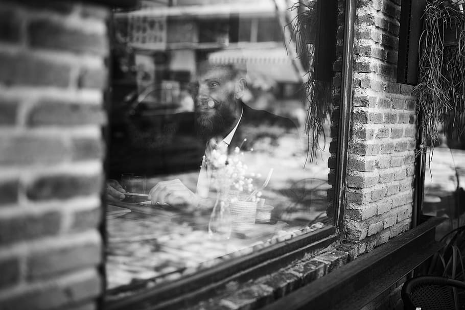 grayscale photo, man, sitting, inside, building, glass window, brick wall, cafe, candid, coffee shop