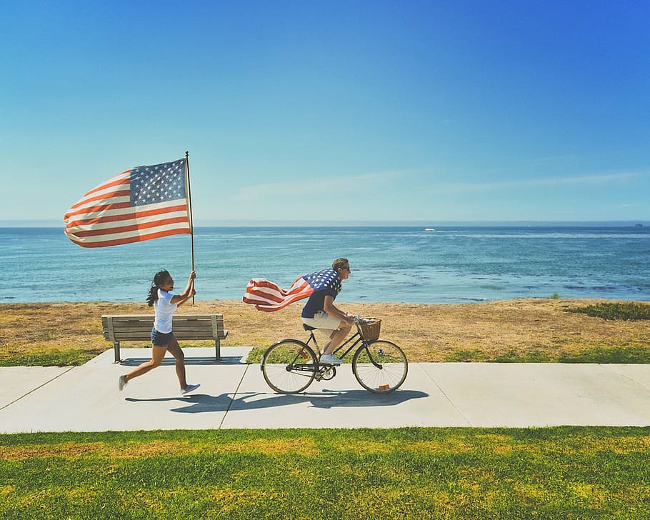 mujer, corriendo, frente, hombre, equitación, bicicleta, sosteniendo, banderas de la bandera de Estados Unidos, al lado, mar