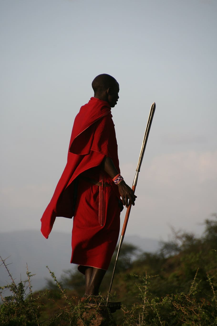 man, wearing, red, top, bottoms, holding, gray, metal spear, standing, rock