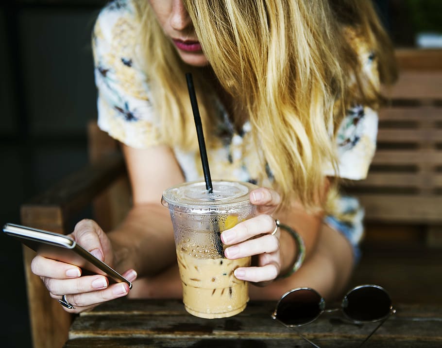 selective, focus photography, blonde, haired woman, sipping, yellow, liquid, using, smartphone, cup
