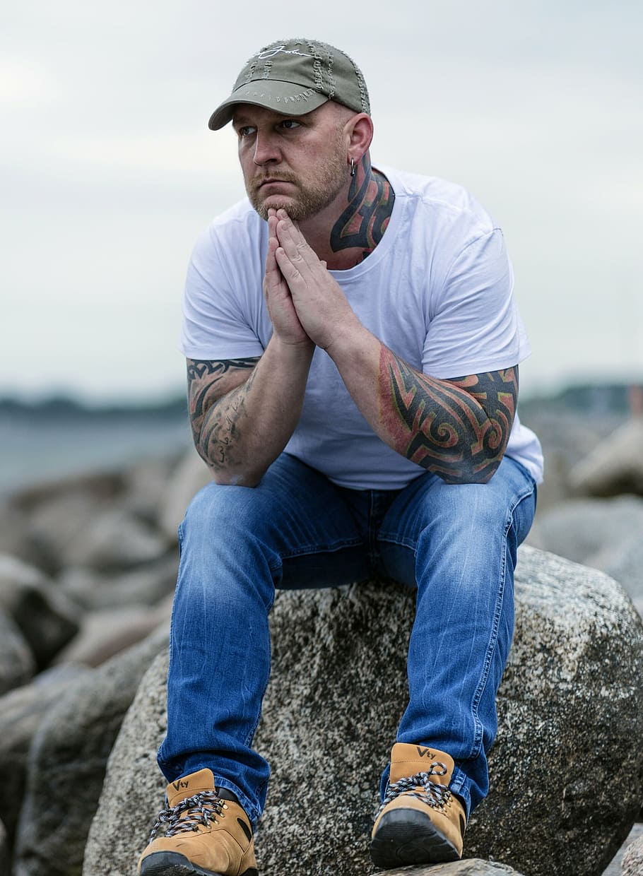 tattooed, man, wearing, gray, baseball cap, white, crew-neck t-shirt, blue, denim jeans, sitting
