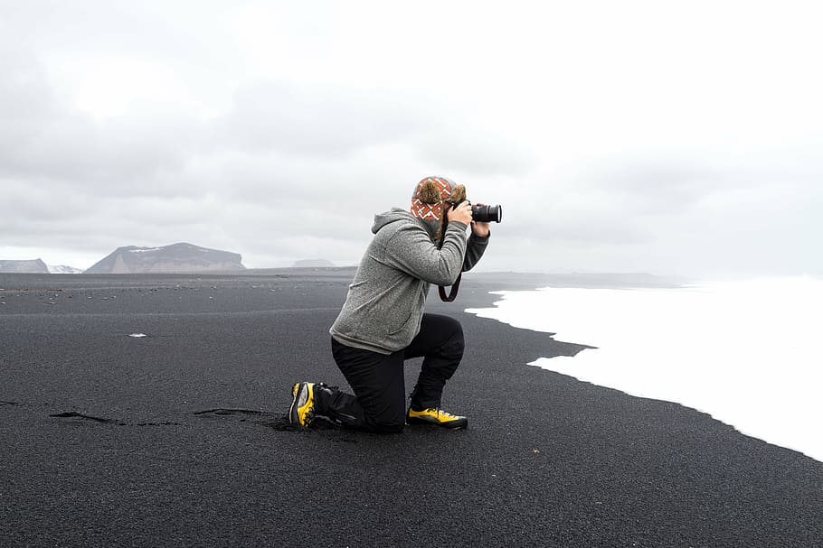hombre, gris, sudadera con capucha, sosteniendo, cámara réflex digital, orilla de la playa, niebla, arrodillado, tomando, foto