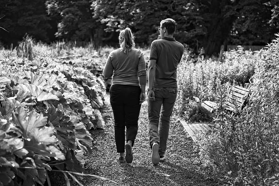 grayscale photography, man, woman, walking, surrounded, plant lot, person, people, side by side, couple