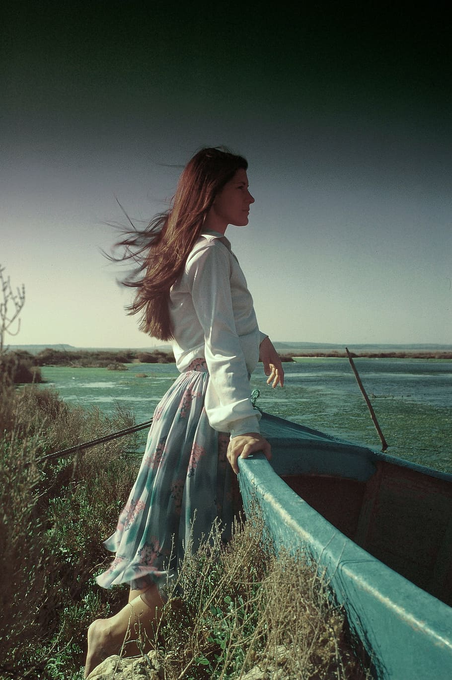 woman, white, shirt, blue, floral, skirt, holding, plastic boat, facing, ocean