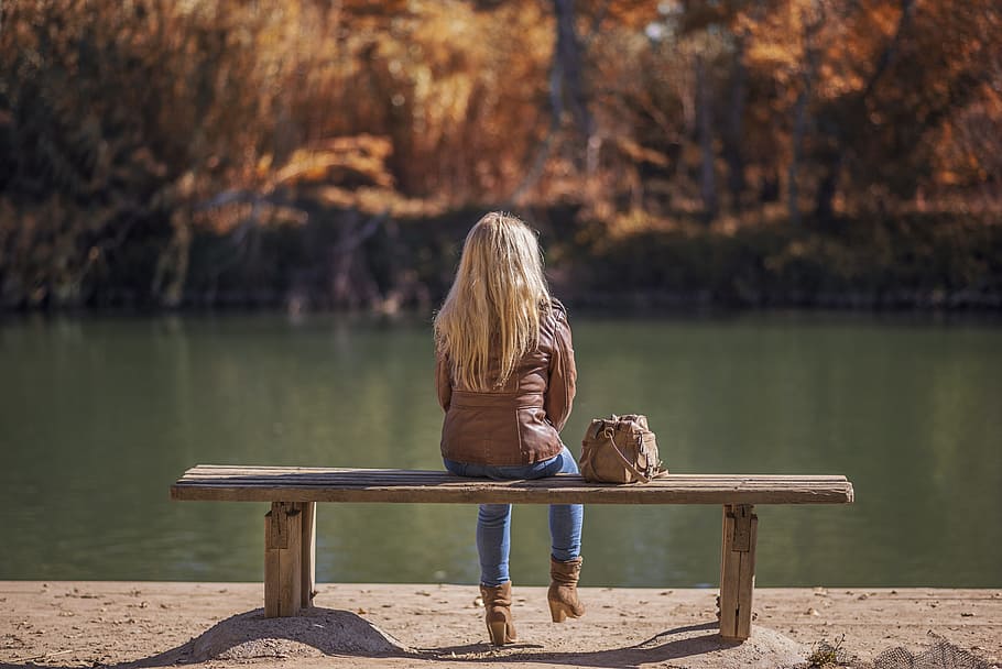 Mujer, vistiendo, marrón, chaqueta de cuero, azul, jeans, sentado, banco, cuerpo de agua, naturaleza