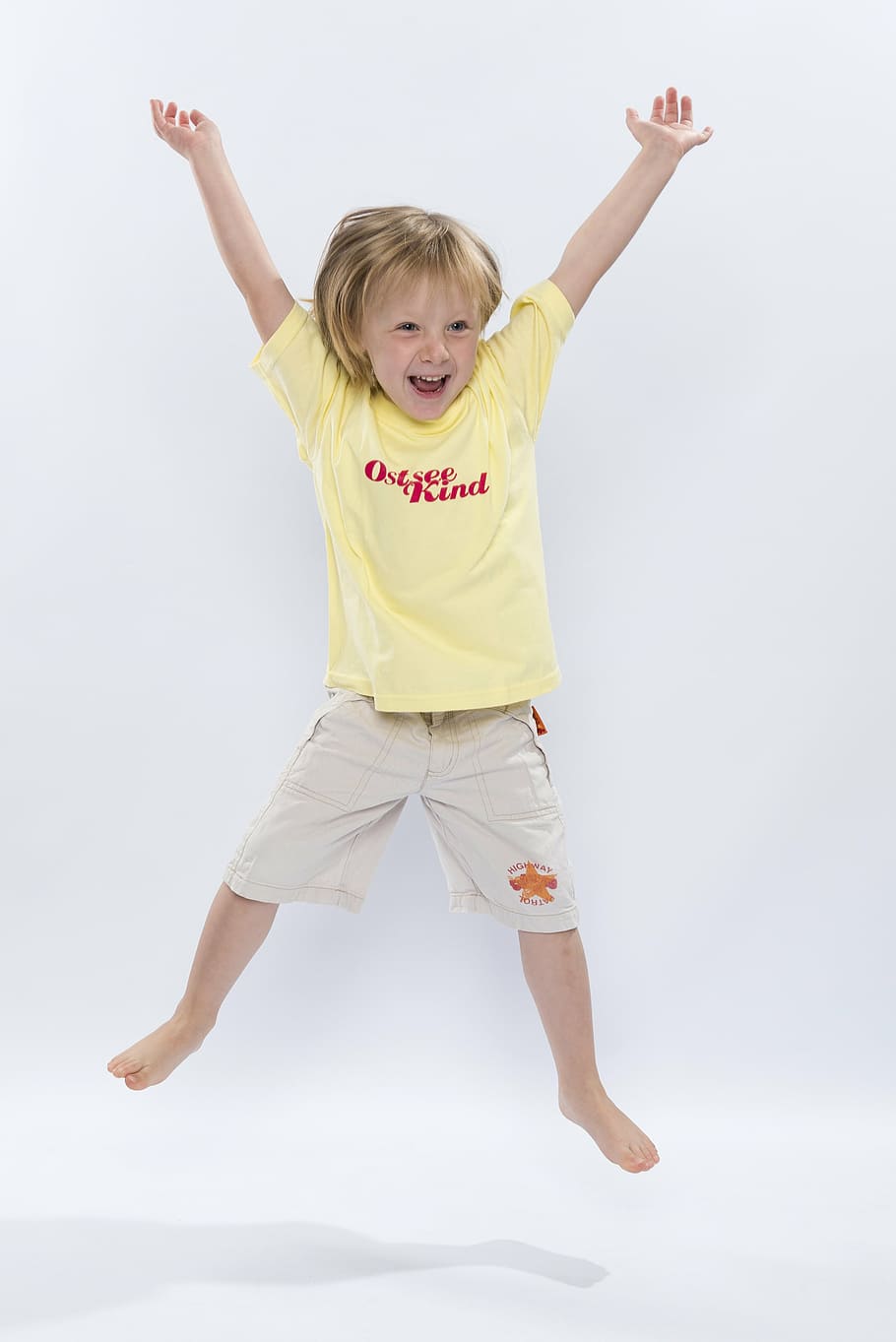 blonde, haired child, wearing, yellow, crew-neck shirt, beige, shorts, child, jumps, laughing