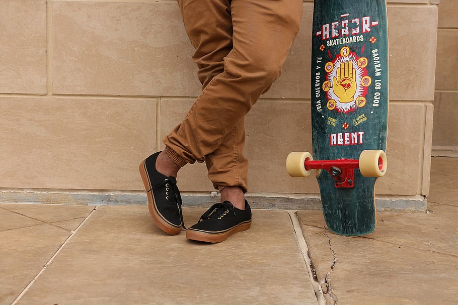 man, holding, longboard, leaning, wall, person, wearing, brown, jeans, pair