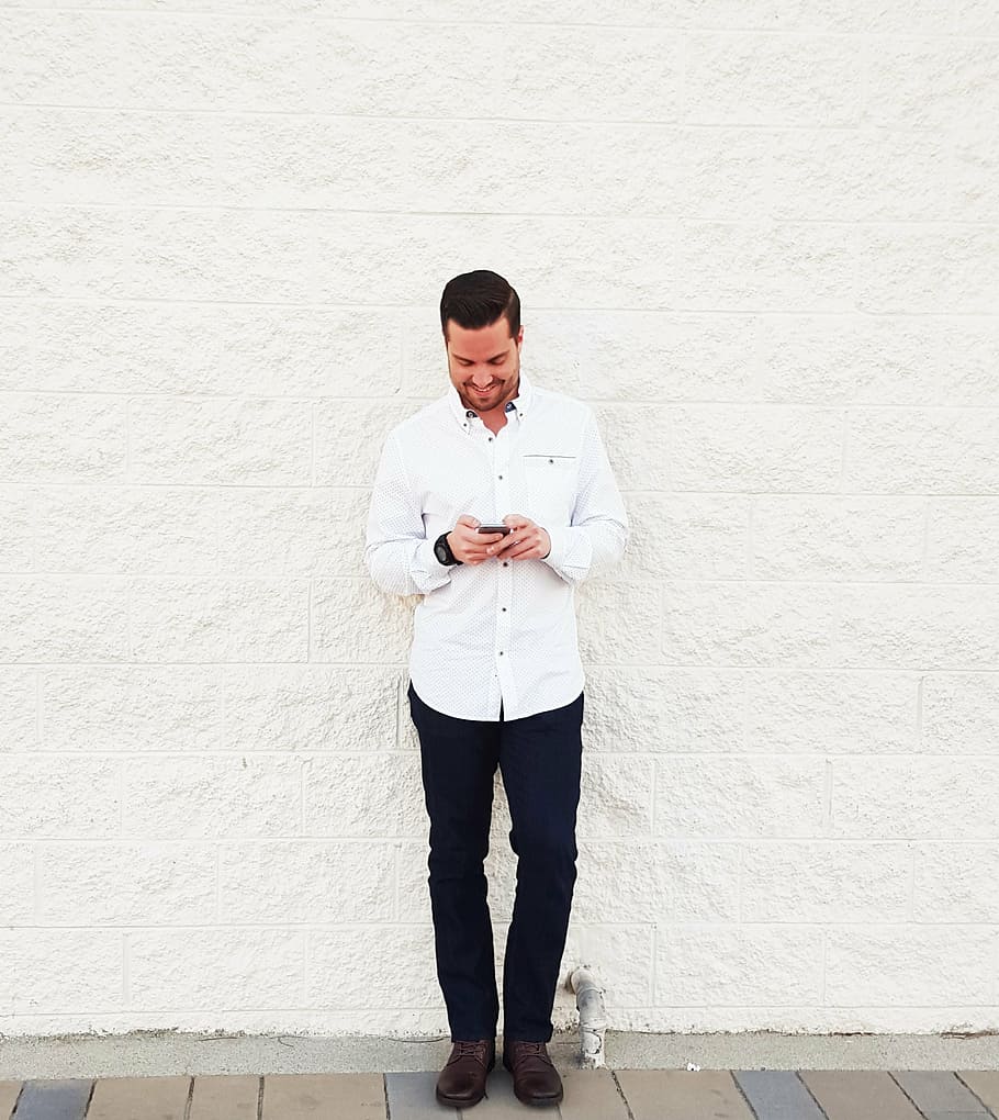 man, wearing, white, dress shirt, black, dress pants, holding, smartphone, leaning, wall