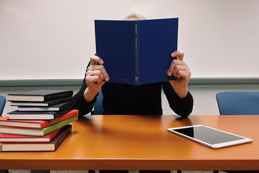 person, holding, blue, book, sitting, infront, brown, wooden, table, read