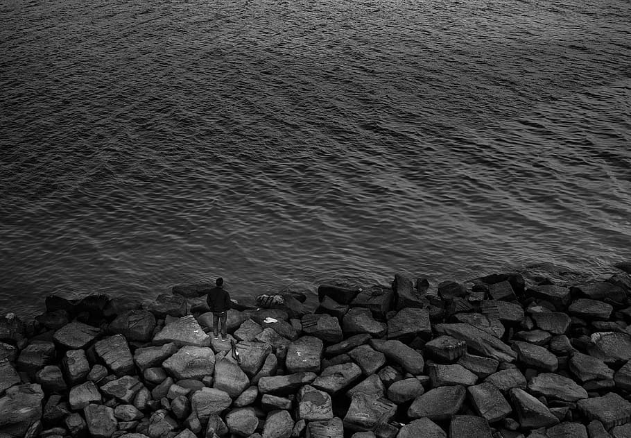 Fotografía en escala de grises, hombre, de pie, rocas, cuerpo, agua, cerca, playa, escala de grises, fotografía