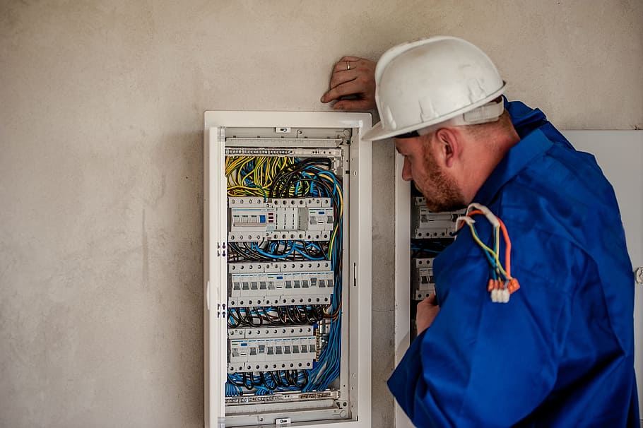 A man in a blue jacket and white hard hat, fixing an electrical panel in a residential home.