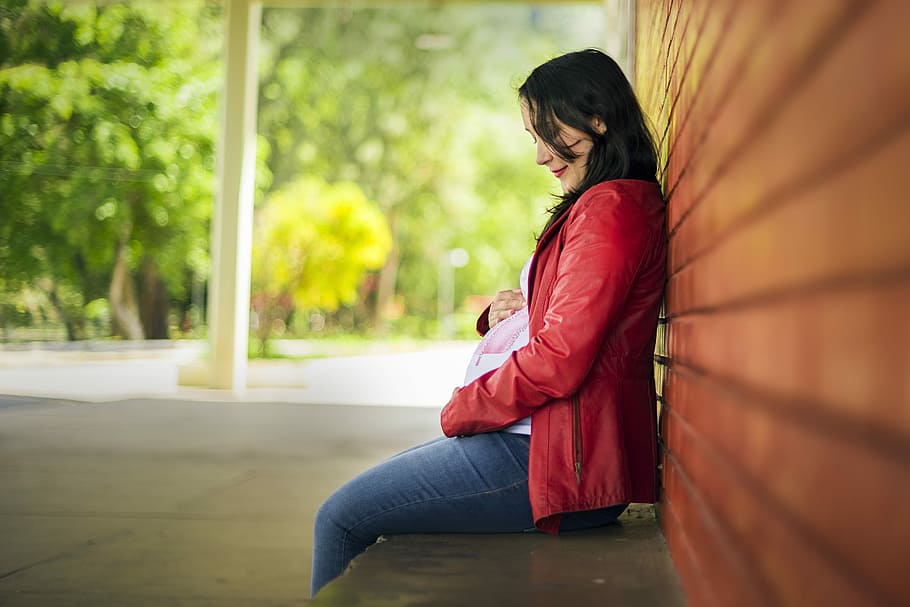 woman, wearing, red, zip-up jacket, sitting, brown, wall, pregnant, pregnant woman, mother
