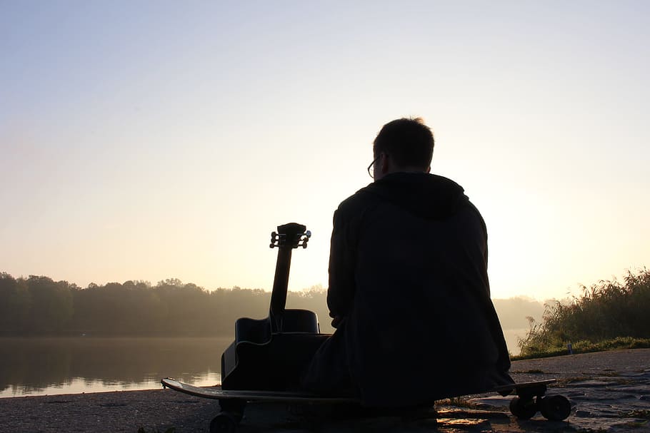 silhouette photo, man, sitting, guitar, facing, lake, across, forest, daytime, musician