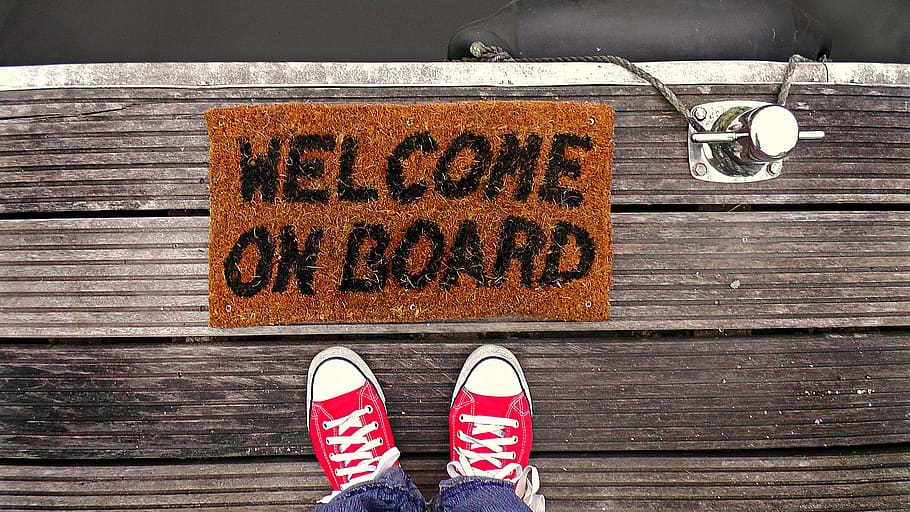 A pair of converse trainers stand on wooden boards, in front of a door mat which says 'welcome on board'. There are silver coloured bolts on the floor next to the mat which imply that this person is standing on a dock. 