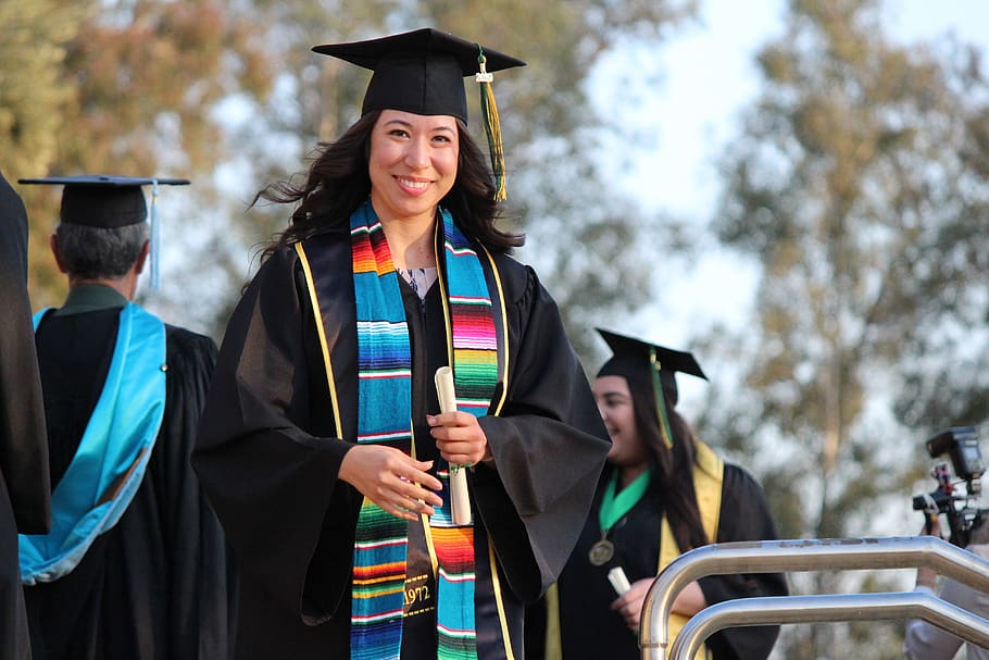 Person dressed in graduation regalia with a paper in her hand and other graduates in the background.