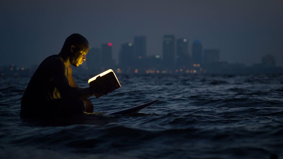 man reading book, sitting, surfboard, person, reading, book, lamp, body, water, background