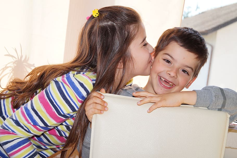 girl kisses, smiling, boy, sitting, gray, leather dining chair, daytime, siblings, sister, good luck brother